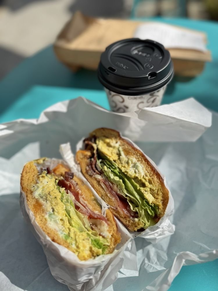 Chicken club bagel sandwich with bacon and lettuce in paper wrap next to a coffee cup