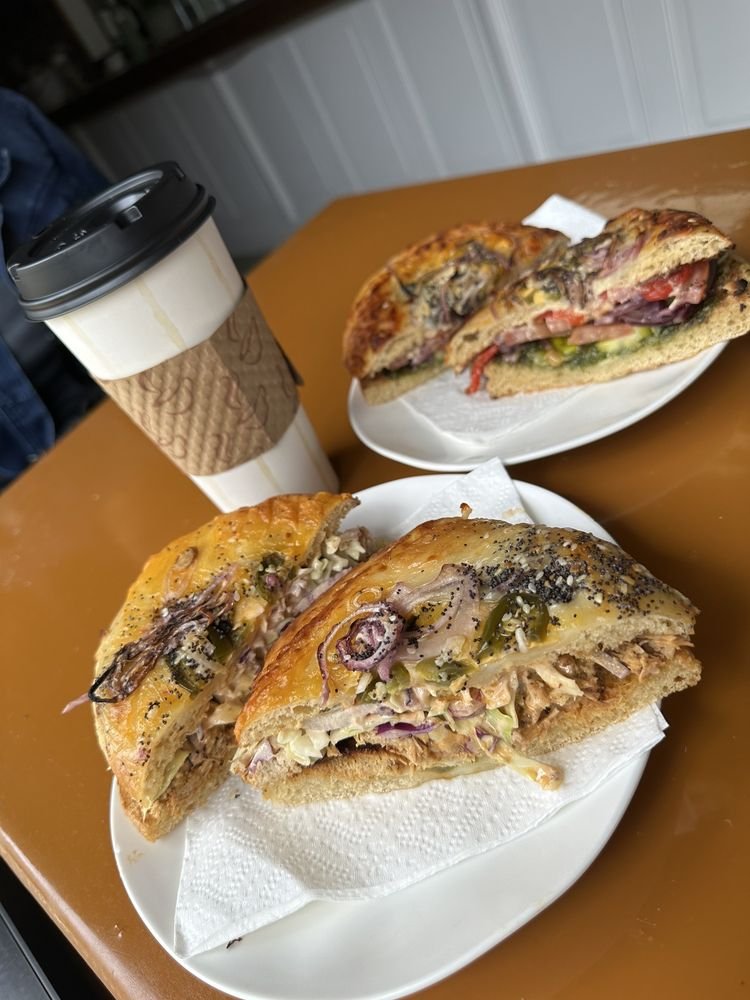 Two bagel sandwiches on plates, one with slaw in front, with a coffee